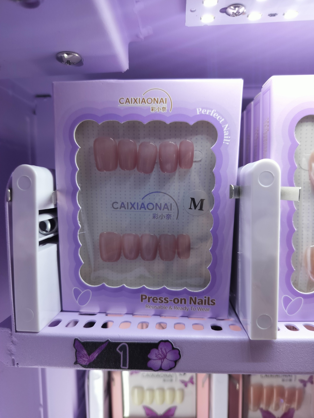 Neutral pink press-on nails in lavender packaging on a vending machine shelf.