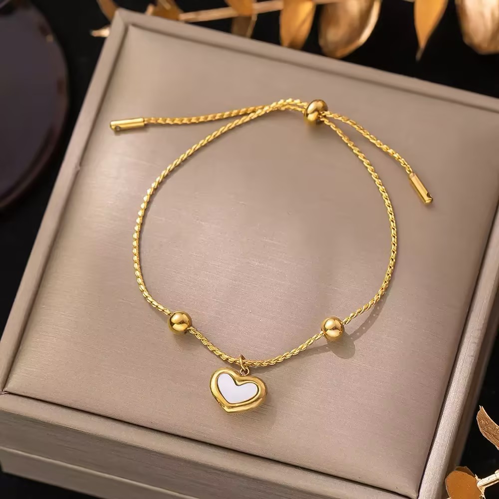 A gold-tone cord-style bracelet with a small white heart centerpiece in a display box.